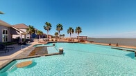 Located at 1514 Ennis Joslin Rd in Corpus Christi, Texas, this single-story waterfront community facility offers an impressive resort-style outdoor experience with direct views of the bay. The centerpiece of the property is a large, freeform swimming pool with crystal-clear turquoise water, accompanied by a separate spa, all set against a backdrop of mature palm trees and open water views stretching to the horizon. A covered pergola provides shaded seating near the water's edge, while lounge chairs and deck seating areas invite relaxation along the generous pool deck, finished in warm-toned concrete and stone tile. The building itself features a striking facade with large, arched and floor-to-ceiling windows that reflect the pool and bay beyond, allowing natural light to flood the interior living areas. A tiled patio runs along the exterior, furnished with wicker seating and bordered by landscaped beds with tropical plantings. Inside, the sun-filled common area showcases tall, multi-pane windows that frame unobstructed views of the pool and waterway, creating a seamless connection between indoor and outdoor spaces. The overall ambiance is coastal and relaxed, with an emphasis on panoramic water views, abundant natural light, and outdoor leisure amenities that define the South Texas waterfront lifestyle.