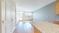 This single-floor unit offers a clean, modern layout designed for comfortable everyday living. The open-concept living and dining area is anchored by warm hardwood-style flooring that flows throughout the main spaces, while large windows fitted with vertical blinds invite abundant natural light and provide views of the surrounding greenery. The living area is spacious and versatile, with softly painted walls in neutral tones that create a calm, welcoming atmosphere.

The kitchen is thoughtfully designed with warm maple cabinetry, granite countertops, and a central island that serves as both a prep space and a natural divider between the kitchen and living areas. White appliances, including a refrigerator, range, microwave, and dishwasher, are neatly integrated, and a light blue tile backsplash adds a subtle pop of color.

The bedroom offers plush wall-to-wall carpeting, a ceiling fan, and a large window that fills the room with natural light, creating a comfortable and restful retreat. The bathroom continues the cohesive design aesthetic with matching maple cabinetry, granite countertops, a large mirror with overhead lighting, and a shower-tub combination, all set on the same warm hardwood-style flooring found throughout the unit.