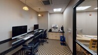 Located at 2500 Clarendon Blvd in Arlington, Virginia, this single-floor business center is designed to meet the productivity needs of guests and visitors. The room features a sleek, built-in desk running along the walls, accommodating multiple iMac computer workstations equipped with keyboards and mice, providing ample space for simultaneous users. Ergonomic mesh-back chairs are positioned at each station, ensuring comfort during extended work sessions. Pendant lighting fixtures hang from the drop-panel ceiling, supplemented by recessed fluorescent lighting to create a well-lit workspace. A dedicated area along one wall houses a multifunction printer and copier atop a storage cabinet, accompanied by a recycling bin, ensuring all essential office needs are met in one place. The floor is covered in a patterned carpet with a geometric motif, adding a touch of visual interest to the otherwise neutral, professionally toned interior. A glass-paneled door with a black frame connects the business center to the broader lobby area, offering a view of the bright, open lounge beyond. The space is compact yet efficiently arranged, making it a convenient and fully equipped stop for travelers requiring reliable workspace access.