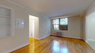 This single-floor residence combines classic architectural details with functional design throughout its thoughtfully laid-out rooms. The living area is a standout, showcasing warm, polished hardwood floors that reflect natural light pouring in through a large window framed by views of mature greenery. Crown molding runs along the ceiling perimeter, lending an elegant, finished look to the space. A classic cast-iron radiator beneath the window adds to the room's timeless character, while a wall-unit air conditioner ensures year-round comfort.

The kitchen is designed as a galley-style space, efficiently lined on both sides with rich, warm-toned wood cabinetry and granite countertops. Black appliances, including a refrigerator, gas range, microwave, and dishwasher, provide a cohesive look, while tile flooring keeps the area practical and easy to maintain. A pass-through counter opens the kitchen toward the living and dining area, enhancing the sense of openness and connectivity between spaces.

The bathroom is clean and functional, featuring tile flooring, a full tub-shower combination with subway tile surround, a wood-toned vanity with an integrated sink, and a window that brings in natural light. Neutral tones throughout the space create a calm, cohesive atmosphere that feels both classic and move-in ready.