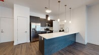 Located at 310 Madison Ave in Elizabeth, NJ, this modern single-floor residence offers a thoughtfully designed open-concept layout with high-end finishes throughout. The kitchen serves as a striking centerpiece, anchored by a large island with a bold blue wood-textured base and a crisp white quartz countertop. Stainless steel appliances — including a French door refrigerator, gas range, and microwave — are complemented by dark espresso cabinetry that extends to the ceiling, providing generous storage. A trio of pendant lights with brushed brass fixtures illuminates the island, adding warmth and style. The kitchen flows seamlessly into a spacious, open living area finished with rich dark hardwood-style flooring, offering a versatile and airy environment. A dedicated in-unit laundry area houses a full-size front-loading washer and dryer, adding everyday convenience. The bathroom is elegantly appointed with large-format marble-look tile on both the floors and tub surround, paired with a double-sink vanity featuring dark wood cabinetry and a clean white countertop. A wide backlit mirror completes the refined look. The overall aesthetic throughout the residence is clean, contemporary, and cohesive, combining modern design elements with functional living spaces.