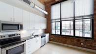 Unit 117 is a single-floor loft-style space that masterfully blends industrial character with modern finishes. The architecture showcases exposed brick walls, warm wood-plank ceilings, and visible ductwork — hallmarks of converted industrial design — creating a distinctive and dynamic aesthetic throughout.

The kitchen is a standout feature, outfitted with sleek white shaker cabinetry extending to the ceiling, dark stone countertops, a subway tile backsplash, and stainless steel appliances including a dishwasher and range. A deep single-basin sink sits beneath the cabinetry, while an industrial-style duct runs overhead, reinforcing the space's warehouse-inspired character. Large, multi-pane black-framed windows line the brick wall, allowing abundant natural light to pour in and offering views of the surrounding streetscape.

A corridor connects the main living areas, with clean white walls contrasting the raw textures of wood and brick visible beyond. The polished concrete-style flooring runs throughout, providing a cohesive and low-maintenance foundation that complements the overall industrial-modern design language. The soaring ceiling height enhances the sense of openness and volume, making this a compelling and characterful space.