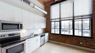 Unit 117 is a single-floor loft-style space that masterfully blends industrial character with modern finishes. The architecture showcases exposed brick walls, warm wood-plank ceilings, and visible ductwork — hallmarks of converted industrial design — creating a distinctive and dynamic aesthetic throughout.

The kitchen is a standout feature, outfitted with sleek white shaker cabinetry extending to the ceiling, dark stone countertops, a subway tile backsplash, and stainless steel appliances including a dishwasher and range. A deep single-basin sink sits beneath the cabinetry, while an industrial-style duct runs overhead, reinforcing the space's warehouse-inspired character. Large, multi-pane black-framed windows line the brick wall, allowing abundant natural light to pour in and offering views of the surrounding streetscape.

A corridor connects the main living areas, with clean white walls contrasting the raw textures of wood and brick visible beyond. The polished concrete-style flooring runs throughout, providing a cohesive and low-maintenance foundation that complements the overall industrial-modern design language. The soaring ceiling height enhances the sense of openness and volume, making this a compelling and characterful space.