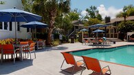 Located at 2225 Ashley River Rd in Charleston, South Carolina, this outdoor amenity space offers a well-appointed resort-style experience designed for relaxation and community enjoyment. The centerpiece is a generously sized swimming pool with clear, blue-green water, framed by a broad concrete sundeck that accommodates ample seating. Vibrant orange lounge chairs and tables with navy blue umbrellas are arranged throughout the deck, providing both shaded and sun-filled areas for residents. Towering palm trees and mature deciduous trees line the perimeter, offering natural shade and a distinctly Southern ambiance. A cabana-style seating area adds an additional retreat for socializing. The single-story clubhouse building, clad in gray horizontal siding with large windows and a covered entrance, anchors one side of the pool area and provides convenient access to interior amenities. Adjacent to the clubhouse, a white-painted building with traditional dark shutters, gridded windows, and manicured shrub landscaping serves as a secondary access point, secured with a keypad entry. The overall atmosphere is lush and inviting, with mature tree canopies filtering natural light across the grounds, creating a tranquil, park-like setting characteristic of the Charleston Lowcountry.