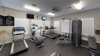 This fitness center is a fully equipped, single-floor facility designed to support a wide range of workout routines. The space features durable rubber flooring throughout, providing safety and comfort during exercise. Natural light floods the room through multiple windows fitted with white blinds, complementing the bright fluorescent lighting and ceiling fans that maintain a comfortable environment.

The cardio zone is well-stocked with Matrix-brand treadmills and elliptical trainers, alongside a rowing machine, all positioned for easy access and optimal use of the floor space. A large wall-mounted mirror runs along one side of the room, visually expanding the space and allowing users to monitor their form. A ballet-style barre is mounted along the mirrored wall, adding a functional element for stretching and balance training.

For strength training, the facility includes a multi-station Matrix cable machine capable of a variety of exercises, as well as an adjustable weight bench. A full rack of hex dumbbells is neatly organized on a tiered stand near the entrance, offering a comprehensive range of free weights. Multiple wall-mounted televisions are positioned throughout for entertainment during workouts, and a wall clock helps users manage their time. A defibrillator is also mounted on the wall, reflecting attention to safety. Entry is through elegant white-framed glass double doors.