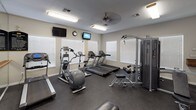 This fitness center is a fully equipped, single-floor facility designed to support a wide range of workout routines. The space features durable rubber flooring throughout, providing safety and comfort during exercise. Natural light floods the room through multiple windows fitted with white blinds, complementing the bright fluorescent lighting and ceiling fans that maintain a comfortable environment.

The cardio zone is well-stocked with Matrix-brand treadmills and elliptical trainers, alongside a rowing machine, all positioned for easy access and optimal use of the floor space. A large wall-mounted mirror runs along one side of the room, visually expanding the space and allowing users to monitor their form. A ballet-style barre is mounted along the mirrored wall, adding a functional element for stretching and balance training.

For strength training, the facility includes a multi-station Matrix cable machine capable of a variety of exercises, as well as an adjustable weight bench. A full rack of hex dumbbells is neatly organized on a tiered stand near the entrance, offering a comprehensive range of free weights. Multiple wall-mounted televisions are positioned throughout for entertainment during workouts, and a wall clock helps users manage their time. A defibrillator is also mounted on the wall, reflecting attention to safety. Entry is through elegant white-framed glass double doors.