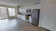 This modern single-floor space offers a clean, open-concept layout designed for both comfort and functionality. The kitchen is a standout feature, showcasing floor-to-ceiling white cabinetry that maximizes storage while maintaining a crisp, minimalist aesthetic. Stainless steel appliances — including a refrigerator, electric range, built-in microwave, and dishwasher — lend a polished, contemporary feel to the cooking area. A single-basin sink is positioned beneath a window, allowing natural light to brighten the workspace throughout the day. The kitchen flows seamlessly into the open living area, creating a spacious, connected environment that feels airy and inviting. Rich, dark hardwood-style flooring runs throughout, providing a warm contrast to the bright white cabinetry and light-toned walls. Recessed lighting in the ceiling offers consistent, well-distributed illumination across the space. A ceiling-mounted HVAC vent ensures year-round climate comfort. The neutral color palette and streamlined design create a versatile backdrop adaptable to a variety of interior styles, making this a well-appointed and thoughtfully designed living space.