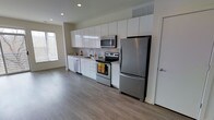 This modern single-floor space offers a clean, open-concept layout designed for both comfort and functionality. The kitchen is a standout feature, showcasing floor-to-ceiling white cabinetry that maximizes storage while maintaining a crisp, minimalist aesthetic. Stainless steel appliances — including a refrigerator, electric range, built-in microwave, and dishwasher — lend a polished, contemporary feel to the cooking area. A single-basin sink is positioned beneath a window, allowing natural light to brighten the workspace throughout the day. The kitchen flows seamlessly into the open living area, creating a spacious, connected environment that feels airy and inviting. Rich, dark hardwood-style flooring runs throughout, providing a warm contrast to the bright white cabinetry and light-toned walls. Recessed lighting in the ceiling offers consistent, well-distributed illumination across the space. A ceiling-mounted HVAC vent ensures year-round climate comfort. The neutral color palette and streamlined design create a versatile backdrop adaptable to a variety of interior styles, making this a well-appointed and thoughtfully designed living space.