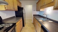 This single-floor residence offers a thoughtfully organized layout with distinct living, sleeping, and kitchen areas. The galley-style kitchen is efficiently designed with warm oak cabinetry lining both walls, dark speckled countertops, a stainless steel sink, and a full suite of appliances including a refrigerator, range, and dishwasher. Tile flooring runs throughout the kitchen, providing a clean and durable surface. Adjacent to the kitchen, the open living area features neutral carpet and leads to a private balcony through a full-width sliding glass door, allowing abundant natural light and outdoor views of surrounding greenery. A separate bedroom offers a comfortable, carpeted retreat with a window that brings in additional natural light. The bathroom is well-equipped with a full tub and shower combination, a toilet, and a wide vanity featuring the same oak cabinetry and dark countertop consistent with the kitchen, complemented by a large mirror and overhead lighting. Throughout the space, neutral tones and clean finishes create a calm, functional atmosphere suitable for everyday living.