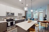 This thoughtfully designed single-floor residence offers a seamless blend of modern aesthetics and functional living. The open-concept layout connects the kitchen and living area, creating a bright, airy atmosphere enhanced by wood-look flooring and recessed lighting throughout.

The kitchen is a standout feature, equipped with a full suite of stainless steel appliances including a refrigerator, range, microwave, and dishwasher. Two-tone cabinetry pairs dark wood lower cabinets with crisp white upper cabinets, complemented by a neutral subway tile backsplash and quartz countertops. A central island with pendant globe lighting provides additional prep space and casual seating with gold-toned bar stools.

The living area is bright and welcoming, featuring large windows and a glass-paned door that frame lush greenery views and invite generous natural light. Transom windows above add to the sense of height and openness.

The bedroom is a calm retreat with large windows overlooking tree-lined surroundings, providing both privacy and natural light. Plush carpeting adds warmth and comfort to the space.

The bathroom features a dark wood vanity with a white quartz countertop and undermount sink, a large mirror, and wood-look flooring. A stacked washer and dryer unit is conveniently integrated into the bathroom, adding in-unit laundry functionality.