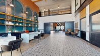 This thoughtfully designed amenity space makes a striking first impression with a soaring double-height lobby entrance. Patterned encaustic-style tile floors transition seamlessly into herringbone wood-look planks, while exposed brick walls, teal arched niches with open shelving, and a bold geometric mural create a vibrant, eclectic aesthetic. A white marble-topped reception desk anchors the lobby area, bathed in abundant natural light from tall glass entry doors and clerestory windows above.

The adjacent dining and lounge area continues the high-ceiling design, featuring a shuffleboard table, bar-height seating, a kitchenette island, and dramatic pendant lighting. Sliding navy barn doors partition the space, offering flexible configurations for gatherings or events.

Hallways are finished with polished concrete floors and house banks of smart package lockers for convenient resident deliveries. A glass-paneled corridor with wood and reeded-glass partitions leads to additional private rooms.

Outdoors, a spacious central courtyard offers lush artificial turf lawn, a modern water feature with stone surround, wooden bench seating, and colorful Adirondack chairs — all framed by the multi-story brick and panel exterior of the building, creating an inviting retreat for residents.
