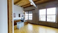 This single-floor loft unit in Grand Rapids' downtown core showcases a striking industrial-chic aesthetic. The open-concept living and kitchen area features soaring ceilings with exposed wood beam construction, visible ductwork, and original structural timber columns. The kitchen includes wood cabinetry, dark countertops, black appliances, and a corrugated metal island. Hardwood floors run throughout the main living areas, while large factory-style windows flood the space with natural light and offer urban cityscape views. A hallway connects private rooms with carpeted floors and closets. The bathroom features tile flooring and a vanity with storage.