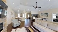 Located at 100 Lake Clair Place in Fayetteville, NC, this single-story space offers a welcoming and well-appointed open-concept interior designed for comfort and functionality. The main living area features soaring vaulted ceilings with recessed lighting and a sleek ceiling fan, creating an airy and expansive atmosphere. Rich, dark hardwood flooring runs throughout, anchoring the space with warmth and sophistication against the soft, neutral wall tones. The generously sized lounge area is furnished with a plush sofa, a patterned accent chair, and a bold coffee table, establishing an inviting gathering space. A kitchen with dark cabinetry, stainless steel appliances, and a breakfast bar is seamlessly integrated into the open floor plan, allowing for easy flow between living and dining functions. Multiple windows allow natural light to pour in from several directions, enhancing the bright and open feel of the interior. A decorative chandelier adds a refined touch near the entryway, complementing the overall modern yet approachable aesthetic. The thoughtful layout and quality finishes make this a versatile and attractive space suited for a variety of uses.