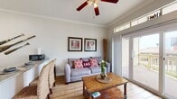 This charming single-floor space is thoughtfully designed with a coastal aesthetic that carries throughout every room. The living area serves as the heart of the unit, featuring vaulted ceilings adorned with crown molding and a ceiling fan, wood-look tile flooring, and an abundance of natural light streaming through a large sliding glass door topped with transom windows that open to an outdoor area. A comfortable gray sofa accented with nautical-themed throw pillows, a rustic wooden coffee table, and decorative bamboo stalks complete the relaxed, beachy ambiance. The kitchen is galley-style, offering ample storage with white cabinetry, granite countertops, and black appliances including a full-size refrigerator and range. Decorative oar wall art adds a playful maritime touch. The bedroom is a serene retreat with vaulted ceilings, a woven headboard, rich navy bedding, coral-motif framed artwork, and a sliding barn door — combining coastal charm with practical design. The bathroom is clean and functional, featuring a granite vanity, a large wall mirror, marble-look tile flooring, and a full tub-shower combination. Together, the rooms create a cohesive, well-appointed space with a warm and inviting coastal character.