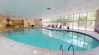 This indoor aquatic and recreation facility centers around a generously sized freeform pool with vivid turquoise water, accessible via stainless steel entry rails and surrounded by a broad, light-toned concrete deck. Recessed ceiling lighting illuminates the pool area evenly, complementing the abundant natural light that pours in through a dramatic floor-to-ceiling glass wall running the length of one side of the space. This expansive window system looks out onto an enclosed outdoor patio area, where mature trees provide a verdant backdrop and additional seating is available.

The poolside deck offers multiple lounge chairs and a seating area with a table and chairs, creating a comfortable space for relaxation. Lush tropical plants, including large yucca specimens, are positioned throughout, adding a resort-like atmosphere to the interior. A dedicated entrance area features a classic wooden balustrade staircase, a bench, and informational signage.

Adjacent to the pool, a separate fitness center is visible through an interior glass partition, equipped with a variety of exercise machines. The facility's light-colored panel walls and drop tile ceiling contribute to a clean, well-maintained aesthetic throughout. Safety equipment is clearly posted, reflecting a commitment to user well-being.