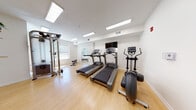 Located in Bloomfield, CT, this single-floor fitness center is thoughtfully designed to support a comprehensive workout experience. The space is divided into distinct functional zones, all unified by light hardwood flooring and crisp white walls that create a clean, energizing atmosphere.

The main cardio area is equipped with multiple Life Fitness treadmills and an elliptical trainer, arranged to face a wall-mounted flat-screen television. Recessed skylight panels and flush ceiling lighting ensure the space is consistently well-lit throughout. A cable resistance machine and free weight rack with a full range of dumbbells provide ample strength training options, while a dedicated weight bench rounds out the resistance training area.

A secondary alcove at the far end of the gym offers a bright, open stretch and functional training zone. Large windows flood this area with abundant natural light and offer views of the surrounding greenery. An elegantly arched full-length mirror is a standout architectural detail, enhancing the sense of openness and allowing users to monitor their form during workouts.

The space connects seamlessly between zones through an interior doorway with a slatted glass partition, maintaining an open yet organized flow. The overall ambiance is bright, practical, and welcoming, making this fitness center a versatile facility suited for a wide range of exercise routines.