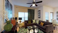 This single-story residence offers a thoughtfully designed open-concept floor plan that connects the living, dining, and kitchen areas into one cohesive, functional space. The living room is anchored by a large sectional sofa, a bold geometric-patterned area rug, and a ceiling fan overhead. Multiple windows fitted with blinds and decorative curtains allow natural light to fill the space, creating a bright and welcoming atmosphere. A glass-top coffee table and decorative wall art add contemporary character throughout.

The dining area sits adjacent to the living room, featuring a round glass dining table with stylish accent chairs and direct access to an exterior door. The kitchen is well-equipped with rich dark wood cabinetry extending to the upper walls, light stone-look countertops, black appliances including a refrigerator, range, and built-in microwave, and a breakfast bar with seating for two.

The bedroom features carpeted flooring, a ceiling fan, large windows, and a spacious walk-in closet outfitted with wire shelving and hanging rods. The bathroom includes a dual-sink vanity with dark wood cabinetry, granite-style countertops, a large mirror, and a tiled tub-shower combination. Wood-look flooring runs throughout the main living areas, tying the design together cohesively.