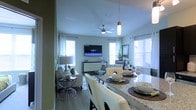 This well-appointed single-floor residence showcases an open-concept design that integrates the dining and living areas into one cohesive, flowing space. The vaulted ceiling adds a sense of height and airiness, while a ceiling fan ensures year-round comfort. Large windows dressed with white curtains and horizontal blinds allow generous amounts of natural light to fill the interior, creating a bright and welcoming ambiance throughout.

The dining area features a granite-topped counter or bar-height surface with seating for several guests, complemented by cream-toned chairs and elegant place settings that lend a polished, refined feel. A stylish pendant light hangs overhead, adding a contemporary touch to the space.

The adjacent living area is thoughtfully arranged with a comfortable seating ensemble, a wall-mounted flat-screen television, and a sleek media console. Decorative accents, including potted greenery and coordinated accessories, contribute to the space's curated aesthetic. The flooring throughout appears to be a smooth hardwood or wood-look material, tying the open layout together with a warm, cohesive finish. Overall, the space balances modern design with functional comfort, making it ideal for both entertaining and everyday living.
