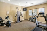 Located in Yucaipa, CA, this single-floor fitness center at 33800 Chapman Heights Rd offers a well-equipped workout space designed for a well-rounded exercise experience. The room features warm, neutral-toned walls complemented by carpet flooring throughout, creating a clean and comfortable environment for physical activity.

The main workout area is outfitted with a variety of exercise equipment, including a treadmill, elliptical trainer, stationary bike, and a multi-function cable strength training machine — providing options for both cardiovascular and resistance training. A wall-mounted flat-screen television is positioned for visibility across the workout floor, making it easy to stay entertained during longer sessions.

Natural light filters into the space through multiple windows fitted with horizontal blinds, allowing for brightness control throughout the day. Recessed ceiling lighting ensures consistent illumination at all hours. A full-length mirror along one wall enhances the sense of space and allows users to monitor their form during exercise.

The entrance area features a glass-paneled door with a security keypad and an interior window looking into an adjacent office or reception area, suggesting a managed community facility. Additional doors provide access to separate rooms within the building. The overall ambiance is practical, tidy, and welcoming — well-suited for regular fitness use.