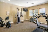 Located in Yucaipa, CA, this single-floor fitness center at 33800 Chapman Heights Rd offers a well-equipped workout space designed for a well-rounded exercise experience. The room features warm, neutral-toned walls complemented by carpet flooring throughout, creating a clean and comfortable environment for physical activity.

The main workout area is outfitted with a variety of exercise equipment, including a treadmill, elliptical trainer, stationary bike, and a multi-function cable strength training machine — providing options for both cardiovascular and resistance training. A wall-mounted flat-screen television is positioned for visibility across the workout floor, making it easy to stay entertained during longer sessions.

Natural light filters into the space through multiple windows fitted with horizontal blinds, allowing for brightness control throughout the day. Recessed ceiling lighting ensures consistent illumination at all hours. A full-length mirror along one wall enhances the sense of space and allows users to monitor their form during exercise.

The entrance area features a glass-paneled door with a security keypad and an interior window looking into an adjacent office or reception area, suggesting a managed community facility. Additional doors provide access to separate rooms within the building. The overall ambiance is practical, tidy, and welcoming — well-suited for regular fitness use.