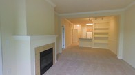 This single-floor space offers an open and functional layout with a seamless flow between the living area and kitchen. The living room is anchored by a tiled fireplace along one wall, providing a warm focal point, while a large built-in shelving unit on the opposite side offers ample display and storage space. Track lighting is mounted along the ceiling, directing light throughout the main area and highlighting the architectural details, including subtle crown molding that frames the upper perimeter of the rooms.

The kitchen is accessible via an open pass-through and features a raised breakfast bar, ideal for casual dining or additional counter space. Warm-toned cabinetry is visible in the background, and a white refrigerator anchors the kitchen area. An arched entryway adds an elegant architectural touch, connecting the kitchen corridor to the rest of the living space.

Soft, neutral carpeting covers the main living area, contributing to a warm and inviting ambiance. A hallway visible near the entry door leads to additional areas of the unit. The overall color palette—cream walls, light carpeting, and white trim—creates a bright and cohesive feel throughout the space.