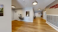 Located at 105 Pomeroy Ave in Meriden, Connecticut, this hallway serves as the welcoming corridor of Pomeroy Place, a thoughtfully designed residential building. The space transitions seamlessly between two distinct flooring materials — warm, light-toned hardwood and a patterned, textured carpet — creating well-defined zones within the corridor.

The entry area features large glass double doors framed by floor-to-ceiling transoms, flooding the space with natural light and providing a clear view to the outdoors. A dark entry mat anchors the threshold, while clean white walls and crisp white baseboards establish a bright, modern aesthetic throughout.

Moving deeper into the hallway, the space opens to reveal a wall-mounted mail center, indicative of a multi-unit residential building, as well as a dual-level water fountain station, ensuring practical accessibility for all residents. Pendant light fixtures with decorative shades provide warm overhead illumination, complementing the recessed and flush-mount ceiling lights found in other areas.

Curated artwork adorns the walls throughout, adding character and a sense of community. A round decorative mirror and a cushioned bench seat provide functional and aesthetic touches in the carpeted lounge-like zone. Multiple doors lead to adjacent spaces, and the overall layout is efficient yet welcoming, reflecting a well-maintained, modern residential environment.