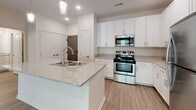 This single-floor unit is designed with a clean, contemporary aesthetic throughout. The kitchen serves as a central highlight, featuring crisp white shaker-style cabinetry extending to the upper walls and a spacious peninsula with granite countertops and an integrated double sink. Stainless steel appliances—including a range, over-the-range microwave, and refrigerator—complement the modern finishes. Recessed and pendant lighting illuminates the space, while light wood-look flooring adds warmth and continuity. A dedicated electrical panel and utility storage are conveniently tucked nearby.

The layout, visible in the overhead scan, reveals an open-concept flow connecting the kitchen to the living area, which benefits from natural light filtering in through a sliding door. The bedroom is carpeted for comfort and features a standard window overlooking a landscaped exterior, paired with a ceiling fan for climate control.

The bathroom maintains the same consistent design language, with a white vanity topped in granite, a large mirror with modern bar lighting, generous upper cabinetry for storage, and wood-look flooring. The overall space presents a cohesive, well-appointed interior with neutral tones and functional finishes throughout.