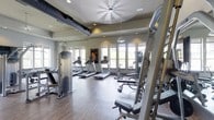 This fitness center offers a thoughtfully designed workout environment that balances functionality with comfort. The open, single-floor layout provides ample room to move freely between stations, creating an inviting atmosphere for exercise. Large windows line the walls, flooding the space with natural light and offering views to the outdoors, helping to create an energizing and airy ambiance. The hardwood-style flooring adds a clean, polished aesthetic throughout. Overhead, a large ceiling fan and recessed lighting work together to maintain comfortable airflow and visibility. Exposed ductwork adds an industrial touch to the otherwise refined interior. The cardio area is outfitted with a row of modern treadmills positioned near the windows, making workouts more engaging. Strength training equipment, including weight benches, cable machines, and multi-function resistance stations, is well-distributed throughout the space. Pendant lighting fixtures complement the room's design, contributing to a warm yet motivating atmosphere. Overall, this fitness center is a comprehensive and well-appointed facility designed to support a wide range of workout routines in a bright, comfortable setting.