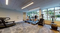 This modern fitness center is thoughtfully designed to accommodate a full range of workout needs within a single, expansive floor. The space is divided into distinct functional zones, seamlessly flowing from one area to the next. The strength training section is generously outfitted with a wide selection of selectorized weight machines, a cable tower, free weight racks stocked with dumbbells and kettlebells, medicine balls, and dedicated bench stations. Large wall-to-wall mirrors amplify the sense of space and allow for proper form monitoring, while wall-mounted flat-screen TVs provide entertainment throughout the workout. The cardio zone features a row of treadmills, elliptical trainers, stationary bikes, and a stair climber, all positioned in front of expansive floor-to-ceiling windows that offer bright natural light and views of a lush, palm tree-lined outdoor setting. TRX suspension training straps are also available for functional fitness. Overhead, recessed linear LED lighting illuminates the entire facility evenly. The floors throughout are covered in durable, neutral-toned carpet tiles that complement the clean, contemporary aesthetic. The overall ambiance is bright, open, and professionally appointed, providing an inviting environment for a complete fitness experience.