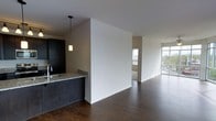 This single-floor space showcases a contemporary open-concept layout with a seamless flow between the kitchen and living areas. The flooring throughout is rich, dark hardwood, lending warmth and contrast to the crisp white walls. The kitchen is positioned along one side, featuring dark cabinetry, a granite countertop peninsula, and a pendant light that adds focused illumination above the workspace. A deep-toned sink and modern faucet complete the functional kitchen design.

A partial wall divides the kitchen from the broader living area, creating a subtle sense of separation while preserving the open feel. Natural light is a defining feature of this space, with large floor-to-ceiling windows visible at the far end of the living area, offering views to an outdoor balcony or terrace. A secondary windowed room is visible through a passage, further enhancing the brightness throughout.

The overall ambiance is clean, modern, and minimalist, with a neutral color palette punctuated by dark tones in the flooring and cabinetry. The layout makes efficient use of space, creating a stylish and functional environment well-suited for contemporary living.