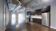 This single-floor space showcases a distinctive industrial aesthetic defined by its exposed ceiling with visible ductwork, steel pipes, and track lighting running across the overhead structure. The open-concept layout connects the living area seamlessly to the kitchen, creating a spacious, loft-like atmosphere.

The kitchen is outfitted with floor-to-ceiling dark espresso cabinetry, offering generous storage. Stainless steel appliances — including a refrigerator, range, built-in microwave, and dishwasher — complement the clean white countertops and a functional single-basin sink. The kitchen's design balances industrial character with modern utility.

The open living area features rich, dark hardwood flooring that extends throughout the space, adding warmth and contrast to the raw ceiling above. A structural column anchors the center of the room, reinforcing the industrial character. Arched windows along the far wall allow natural light to fill the space, brightening the otherwise dramatic interior. The combination of exposed infrastructure, modern finishes, and open layout gives this space a bold, contemporary urban character.