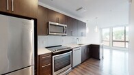 This single-floor residence showcases a thoughtfully designed contemporary interior with high-quality finishes throughout. The kitchen is a standout feature, equipped with full-height dark espresso cabinetry, white quartz countertops, a subway tile backsplash, and a complete suite of stainless steel appliances including a refrigerator, electric range, built-in microwave, and dishwasher. Elegant glass pendant lights hang above the kitchen peninsula, which opens directly to the living area, creating a seamless and functional layout. The living space features light-toned luxury vinyl plank flooring and is bathed in natural light from large windows.

A hallway connects the main living areas to the bedroom, which is finished with plush wall-to-wall carpeting and a window offering outdoor views. The bathroom is finished with large-format marble-look porcelain tile flooring, a dark wood vanity with a white countertop, a framed mirror, and a window for natural light. A dedicated laundry closet houses a stacked washer and dryer unit, adding practical convenience. A sliding barn-style door and recessed lighting throughout complete the modern aesthetic of this well-appointed space.