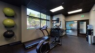 This single-level fitness center on NW Pettygrove Street in Portland's Northwest District offers a comprehensive workout environment designed with a modern, functional aesthetic. The space features a dark grid ceiling with recessed panel lighting that provides consistent illumination throughout, complemented by large windows along one wall that allow natural light to flow in while offering views of the surrounding streetscape.

The main workout area is equipped with a range of cardio machines, including treadmills and elliptical trainers by SportsArt, as well as a cable resistance machine and a seated rowing machine. A dedicated free weights section includes a full dumbbell rack and stability balls for functional training. Rubber flooring throughout provides durability and traction suitable for a variety of exercise activities.

A wall-mounted flat-screen television is positioned for visibility from the cardio equipment. Full-length mirrors along one section of the wall enhance the sense of space and support form-checking during workouts. A water cooler station is available near the entrance area, accompanied by a waste receptacle. The entryway features frosted-glass panel doors, providing a clean and professional transition into the facility. Fitness center rules are posted near the entrance for user reference.
