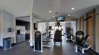 This fitness center offers a comprehensive workout environment designed to accommodate a range of exercise routines. The space features vaulted ceilings that lend an open, airy feel, complemented by recessed lighting that ensures the area is well-lit throughout. The flooring consists of durable rubber gym matting, providing both safety and comfort during workouts.

The equipment selection is extensive, including treadmills, elliptical trainers, stair climbers, and a rowing machine, along with strength training equipment such as a cable machine. A wall-mounted flat-screen television is positioned prominently, allowing members to stay entertained during cardio sessions. Structural support columns are integrated into the layout, and the equipment is arranged to maximize usable floor space while maintaining clear pathways between machines.

A small kitchenette area is situated along one wall, featuring dark cabinetry, a sink, and a full-size refrigerator, offering a practical space for hydration and storage needs. The overall ambiance is clean, functional, and purposefully organized, making this fitness center a well-rounded facility suited for daily exercise.