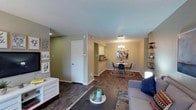 This single-story unit offers a thoughtfully arranged layout that connects its primary living spaces in a cohesive, functional flow. The living room features dark wood-look flooring, a textured ceiling, and neutral warm-toned walls, creating a welcoming atmosphere. The space accommodates a comfortable seating arrangement and opens directly into a dedicated dining area, which is highlighted by a modern chandelier, matching wood-look flooring, and room for a round table with seating for four. Adjacent to the dining area, the kitchen is equipped with white cabinetry, dark countertops, black appliances including a refrigerator and electric range, and a decorative tile backsplash that adds visual interest.

The unit includes two bedrooms, both finished with plush carpeting and ceiling fans for comfort. One bedroom features an en-suite vanity area with a large mirror, white cabinetry, and dark countertops, alongside a separate walk-in closet. The second bedroom offers a window with natural light and ample room for furnishings. The bathroom includes dark wood-look flooring, a vanity with dark countertops, white cabinetry, and a shower-tub combination. Throughout the unit, the design maintains a consistent, understated aesthetic.
