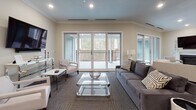 This single-floor space in South Easton, Massachusetts, is thoughtfully designed to serve as a versatile community or gathering area. The layout flows seamlessly between a fully equipped kitchen and multiple distinct lounge and seating zones, all unified by neutral warm tones, crown molding, and recessed lighting throughout.

The kitchen area is a standout feature, showcasing white shaker-style cabinetry, stainless steel appliances including a full-size refrigerator and wall oven, a undermount sink, and a generous island with a white quartz countertop and bar seating for several guests. Pass-through windows connect this area to adjacent spaces, enhancing the open, communicative feel of the floor plan.

The lounge areas are generously sized and well-furnished, offering a variety of seating arrangements suited for both relaxed gatherings and more intimate conversations. One area includes a classic white fireplace mantle, adding warmth and elegance. Wall-mounted televisions are present in multiple zones for added functionality.

Large sliding glass doors open to an outdoor balcony with wrought iron railings, bringing in abundant natural light and views of the surrounding wooded landscape. A dining area with a glass round table and white chairs adds a refined touch. Throughout, plush carpet flooring and crown molding contribute to the space's polished, welcoming ambiance.