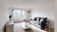 This thoughtfully laid-out one-bedroom, one-bathroom unit offers comfortable single-floor living across a well-proportioned floor plan. The living room serves as the heart of the unit, featuring plush carpeting, a large picture window with outdoor tree views, a wall-mounted AC unit, and baseboard heating. The open sightline from the living area extends into a dedicated dining space, creating a cohesive and functional common area with warm recessed lighting overhead. The galley-style kitchen is efficiently designed with white cabinetry, light countertops, a stainless steel sink, and hardwood-style flooring, providing ample storage and workspace along both sides. The bedroom is generously sized with soft carpeting, a wide window that invites natural light, and baseboard heating, offering a calm and restful environment. The full bathroom includes a wood-toned vanity with a wide countertop, a large mirror with built-in shelving, a combined tub and shower, and vinyl flooring. Throughout the unit, the clean white walls and neutral tones create a bright, airy atmosphere well-suited for a range of interior styles.