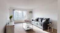 This thoughtfully laid-out one-bedroom, one-bathroom unit offers comfortable single-floor living across a well-proportioned floor plan. The living room serves as the heart of the unit, featuring plush carpeting, a large picture window with outdoor tree views, a wall-mounted AC unit, and baseboard heating. The open sightline from the living area extends into a dedicated dining space, creating a cohesive and functional common area with warm recessed lighting overhead. The galley-style kitchen is efficiently designed with white cabinetry, light countertops, a stainless steel sink, and hardwood-style flooring, providing ample storage and workspace along both sides. The bedroom is generously sized with soft carpeting, a wide window that invites natural light, and baseboard heating, offering a calm and restful environment. The full bathroom includes a wood-toned vanity with a wide countertop, a large mirror with built-in shelving, a combined tub and shower, and vinyl flooring. Throughout the unit, the clean white walls and neutral tones create a bright, airy atmosphere well-suited for a range of interior styles.