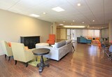Located at 3901 Indian School Rd NE, this single-floor space offers a welcoming and functional layout designed for comfort and collaboration. The interiors are unified by rich hardwood-style flooring and drop tile ceilings with recessed and panel lighting throughout.

The main lounge area is generously sized, featuring a plush gray sectional sofa, vibrant mustard and orange accent chairs, a tufted ottoman, and a decorative dark wood cabinet — all arranged atop a colorful area rug. Large windows with vertical blinds allow natural light to fill the room, creating an open and inviting atmosphere.

A second seating area showcases a teal sectional sofa paired with patterned ottomans, additional accent chairs, and a console table against a soft blue accent wall, lending a distinct and relaxed character to the space.

A dining area includes a round dark table with a mix of modern molded chairs in orange and gray, positioned near a structural column that adds an architectural element to the open floor plan. Adjacent to this area, a compact kitchenette features wood cabinetry and appliances, providing practical convenience.

Wide hallways connect multiple rooms with white panel doors, offering flexible use of the space. The overall design balances functional utility with a contemporary, colorful aesthetic suitable for a variety of professional or community-oriented uses.