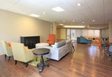 Located at 3901 Indian School Rd NE, this single-floor space offers a welcoming and functional layout designed for comfort and collaboration. The interiors are unified by rich hardwood-style flooring and drop tile ceilings with recessed and panel lighting throughout.

The main lounge area is generously sized, featuring a plush gray sectional sofa, vibrant mustard and orange accent chairs, a tufted ottoman, and a decorative dark wood cabinet — all arranged atop a colorful area rug. Large windows with vertical blinds allow natural light to fill the room, creating an open and inviting atmosphere.

A second seating area showcases a teal sectional sofa paired with patterned ottomans, additional accent chairs, and a console table against a soft blue accent wall, lending a distinct and relaxed character to the space.

A dining area includes a round dark table with a mix of modern molded chairs in orange and gray, positioned near a structural column that adds an architectural element to the open floor plan. Adjacent to this area, a compact kitchenette features wood cabinetry and appliances, providing practical convenience.

Wide hallways connect multiple rooms with white panel doors, offering flexible use of the space. The overall design balances functional utility with a contemporary, colorful aesthetic suitable for a variety of professional or community-oriented uses.