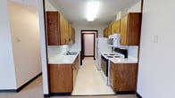 This single-floor space centers around a classic galley-style kitchen designed for efficient use of space. The kitchen is laid out with cabinetry lining both walls, finished in warm honey-toned oak with dark hardware accents that add a traditional character. Upper and lower cabinets provide generous storage on each side, maximizing functionality within the narrow corridor layout.

White laminate countertops run along both sides of the kitchen, offering practical workspace. On one side, a stainless single-basin sink is set beneath the upper cabinetry, accompanied by a textured tile backsplash that adds subtle visual interest. The opposite wall houses a white electric coil-top range and oven, complemented by an over-the-range microwave. A white refrigerator anchors the far end of the kitchen, positioned near a doorway that leads to an adjacent room or hallway.

The flooring throughout the kitchen is light-toned vinyl, easy to maintain and consistent in its clean appearance. Overhead, a flush-mount fluorescent light fixture provides even illumination across the space. The overall ambiance is practical and straightforward, with a functional layout well-suited for everyday use.