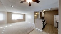 This single-floor residential space offers a practical and comfortable layout designed for everyday living. The open-concept floor plan seamlessly connects the living area to the kitchen, creating a cohesive and functional environment. The living area is finished with plush carpeting in a neutral beige tone, contributing to a warm and inviting atmosphere. A ceiling fan with a light fixture is centrally mounted, providing both airflow and ambient illumination. A large window allows natural light to fill the room, enhancing the sense of openness.

The kitchen is positioned at the edge of the living space, separated by a peninsular counter that serves as both a workspace and a casual divider between the two areas. Dark-stained wood cabinetry overhead contrasts with the lighter countertops and beige walls, adding visual depth. The kitchen is equipped with under-counter storage and appliances, with tile flooring that transitions cleanly from the carpeted living area. Recessed lighting above the kitchen provides focused task illumination. The overall color palette of warm beige and neutral tones throughout the space creates a cohesive and move-in ready environment.