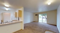 This single-floor residence offers a practical and open layout designed to maximize usable living space. The living area is carpeted and generously sized, creating a comfortable and welcoming atmosphere. A sliding glass door at the far end of the living room floods the space with natural light and provides direct access to an outdoor green area, offering pleasant views of lush landscaping.

The kitchen is positioned adjacent to the living area and is separated by a partial wall with a pass-through counter, maintaining an open and connected feel throughout. Light wood cabinetry lines the upper portion of the kitchen walls, offering ample storage, while a countertop provides functional workspace. The kitchen and entryway feature hard-surface flooring, contrasting with the carpeted living space.

A wall-mounted climate control unit is installed in the living area, providing efficient temperature regulation. The interiors are finished in neutral tones throughout — from the off-white walls to the beige carpeting — giving the space a clean, understated aesthetic that is easy to personalize. Overall, the layout is straightforward and functional, well-suited for everyday living.