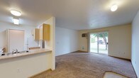 This single-floor residence offers a practical and open layout designed to maximize usable living space. The living area is carpeted and generously sized, creating a comfortable and welcoming atmosphere. A sliding glass door at the far end of the living room floods the space with natural light and provides direct access to an outdoor green area, offering pleasant views of lush landscaping.

The kitchen is positioned adjacent to the living area and is separated by a partial wall with a pass-through counter, maintaining an open and connected feel throughout. Light wood cabinetry lines the upper portion of the kitchen walls, offering ample storage, while a countertop provides functional workspace. The kitchen and entryway feature hard-surface flooring, contrasting with the carpeted living space.

A wall-mounted climate control unit is installed in the living area, providing efficient temperature regulation. The interiors are finished in neutral tones throughout — from the off-white walls to the beige carpeting — giving the space a clean, understated aesthetic that is easy to personalize. Overall, the layout is straightforward and functional, well-suited for everyday living.