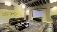 This single-floor space is purpose-built for an immersive cinematic experience. The centerpiece of the room is a large wall-mounted projection screen paired with a ceiling-mounted projector, offering a true theater-quality viewing setup. A sleek media console positioned beneath the screen provides organized storage for entertainment components, complemented by surround sound speakers strategically placed throughout the room.

The seating area features a generous dark leather sectional sofa arranged around a substantial rectangular coffee table, creating a comfortable and stylish viewing environment. The flooring consists of patterned carpet tiles in muted, earthy tones that add texture and warmth to the space while aiding in sound absorption.

Architecturally, the room stands out with its exposed dark beam ceiling, which lends an industrial yet refined character to the space. A hanging drum pendant light adds a soft ambient glow, while wall-mounted accent lighting and decorative circular wall art contribute to the room's polished aesthetic. Warm yellow-toned walls enhance the cozy, intimate atmosphere ideal for movie viewing. A recessed entryway leads into the space, hinting at a thoughtfully designed layout that keeps the theater environment visually and acoustically focused.