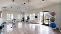 Located at 16755 W Lake Houston Parkway in Houston, TX, this single-room fitness studio is purpose-built for cycling and group exercise. The space features light hardwood-style flooring that extends across the entire room, creating a clean, open workout environment. Vaulted ceilings lend an airy, spacious feel, while track lighting and ceiling fans provide both functional illumination and air circulation throughout.

Multiple Life Cycle spin bikes are arranged across the floor, offering ample space between each station for comfortable use. A full mirrored wall along one side expands the sense of space visually and supports proper form during workouts. A mounted flat-screen display adjacent to the mirrors enables on-demand or instructor-led cycling classes.

Natural light fills the room through several large windows fitted with blinds, and a glass-panel door opens directly to the outdoors, connecting the studio to the surrounding exterior. Stability balls in multiple sizes are stored along the mirrored wall, and step platforms are neatly stacked for additional workout versatility. Wall-mounted accessories and posted guidelines further support a well-organized fitness environment. The bright, white walls and clean design aesthetic contribute to an energizing and motivating atmosphere ideal for group fitness and individual training sessions.