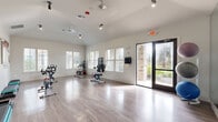 Located at 16755 W Lake Houston Parkway in Houston, TX, this single-room fitness studio is purpose-built for cycling and group exercise. The space features light hardwood-style flooring that extends across the entire room, creating a clean, open workout environment. Vaulted ceilings lend an airy, spacious feel, while track lighting and ceiling fans provide both functional illumination and air circulation throughout.

Multiple Life Cycle spin bikes are arranged across the floor, offering ample space between each station for comfortable use. A full mirrored wall along one side expands the sense of space visually and supports proper form during workouts. A mounted flat-screen display adjacent to the mirrors enables on-demand or instructor-led cycling classes.

Natural light fills the room through several large windows fitted with blinds, and a glass-panel door opens directly to the outdoors, connecting the studio to the surrounding exterior. Stability balls in multiple sizes are stored along the mirrored wall, and step platforms are neatly stacked for additional workout versatility. Wall-mounted accessories and posted guidelines further support a well-organized fitness environment. The bright, white walls and clean design aesthetic contribute to an energizing and motivating atmosphere ideal for group fitness and individual training sessions.