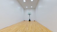 This dedicated indoor sports court offers an impressive recreational space designed for serious athletic use. The court is defined by its gleaming, light-toned hardwood flooring that runs the full length of the room, providing a professional-grade surface well-suited for basketball, racquetball, or a variety of other activities.

The space features notably high ceilings that allow for full, unobstructed play, while the clean white walls on all sides create a bright, open atmosphere. Recessed skylight-style ceiling fixtures provide consistent, even illumination throughout the court, ensuring excellent visibility during any activity.

Two wall-mounted basketball backboards and hoops are positioned at opposite ends of the court, complemented by a portable adjustable basketball hoop stationed at center court. A set of high-placed windows along one wall introduces natural light and provides a glimpse of the exterior surroundings, adding to the airy feel of the space.

A single access door is discreetly integrated into one of the side walls for convenient entry and exit. The overall design is clean, functional, and purposefully minimal — prioritizing performance and versatility. This private indoor court represents a rare and valuable amenity, offering a dedicated environment for year-round athletic training and recreation.