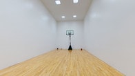 This dedicated indoor sports court offers an impressive recreational space designed for serious athletic use. The court is defined by its gleaming, light-toned hardwood flooring that runs the full length of the room, providing a professional-grade surface well-suited for basketball, racquetball, or a variety of other activities.

The space features notably high ceilings that allow for full, unobstructed play, while the clean white walls on all sides create a bright, open atmosphere. Recessed skylight-style ceiling fixtures provide consistent, even illumination throughout the court, ensuring excellent visibility during any activity.

Two wall-mounted basketball backboards and hoops are positioned at opposite ends of the court, complemented by a portable adjustable basketball hoop stationed at center court. A set of high-placed windows along one wall introduces natural light and provides a glimpse of the exterior surroundings, adding to the airy feel of the space.

A single access door is discreetly integrated into one of the side walls for convenient entry and exit. The overall design is clean, functional, and purposefully minimal — prioritizing performance and versatility. This private indoor court represents a rare and valuable amenity, offering a dedicated environment for year-round athletic training and recreation.