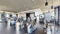 Located at 310 Broad Street in the heart of Charleston, South Carolina, this contemporary fitness center offers a generously sized, single-level exercise space designed to support a comprehensive workout experience. The facility is thoughtfully laid out across an open floor plan, with distinct zones dedicated to cardio training, strength machines, and free weights.

The cardio area features a full lineup of modern equipment including treadmills, ellipticals, stair climbers, and stationary bikes, all arranged along large windows that flood the space with natural light and provide views of the surrounding Charleston skyline. A bold orange accent ceiling distinguishes this zone and adds an energetic visual character, complemented by sleek pendant lighting.

The strength and free-weight section is anchored by a dramatic full-wall mural depicting a serene South Carolina lowcountry marsh landscape, creating a calming contrast to the active environment. Wall-mounted flat-screen televisions are positioned throughout for entertainment during workouts. Rubberized flooring provides both safety and durability across the exercise areas.

The overall design balances modern aesthetics with functional layout, using dark accent walls, recessed and pendant lighting, and floor-to-ceiling windows to create an inviting, well-lit atmosphere suited for a wide range of fitness activities.